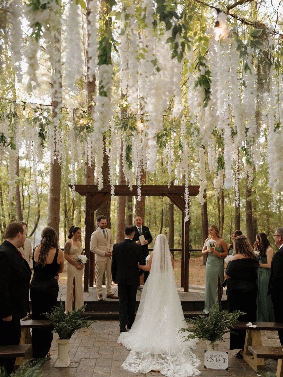 Wedding couple standing under a wooden arbor with hanging white flowers in a forest