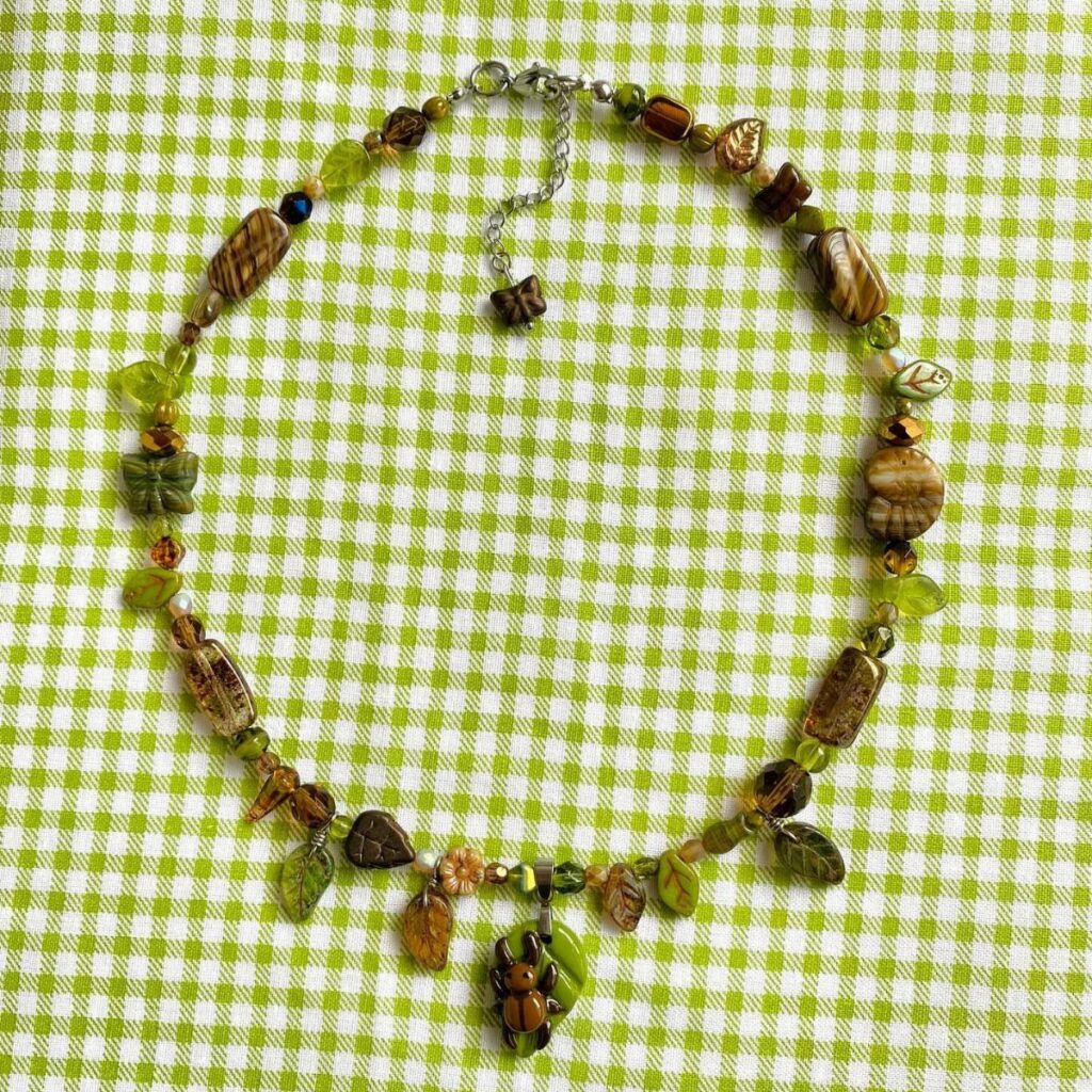 Handmade beaded necklace with green leaf and insect charms on checkered fabric