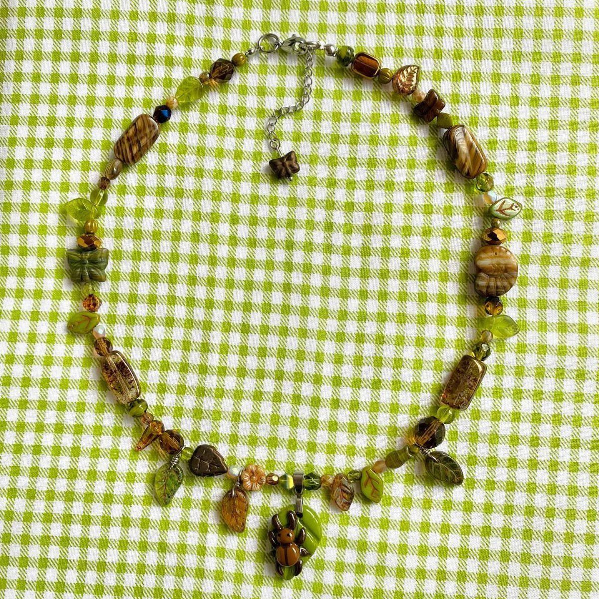 Handmade beaded necklace with green leaf and insect charms on checkered fabric