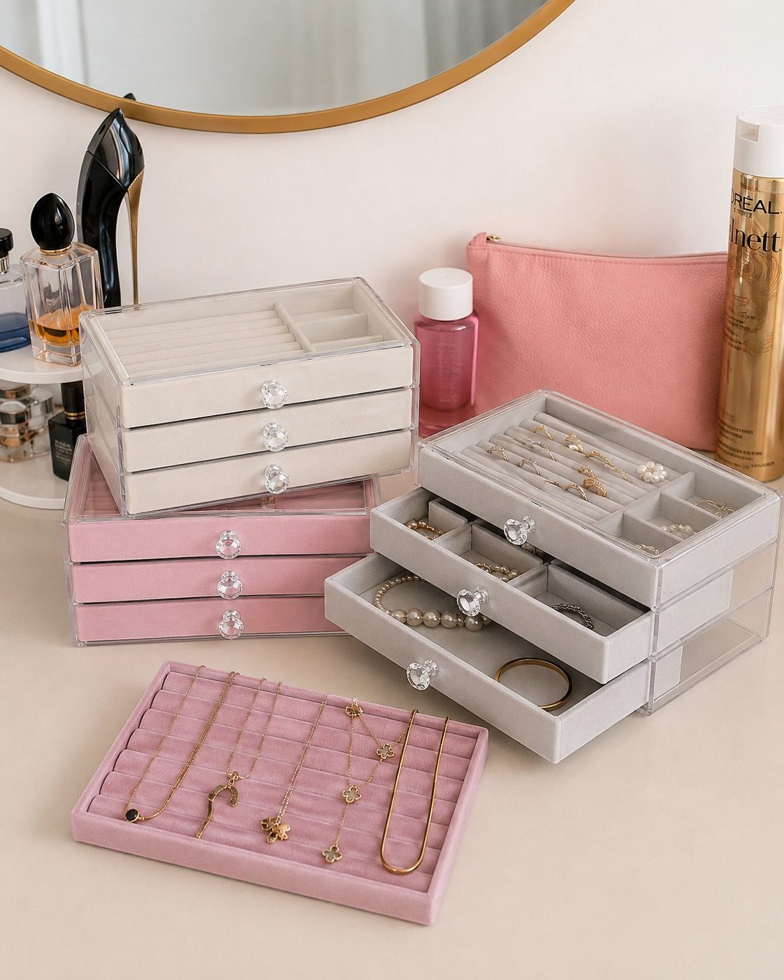Velvet-lined acrylic jewelry boxes in pink, white, and grey holding various necklaces and rings.