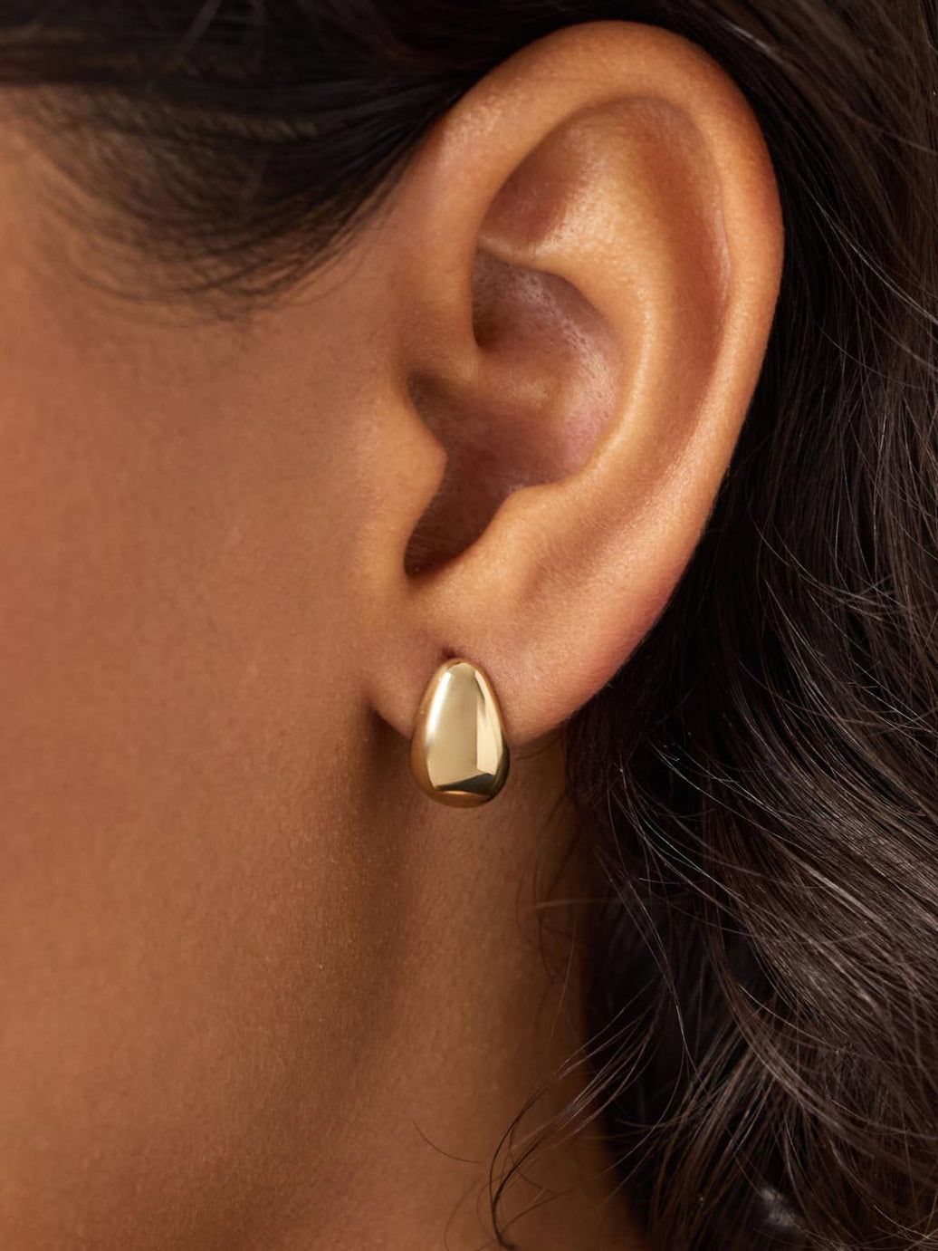 Gold stud earring with a smooth organic shape worn on a person's ear