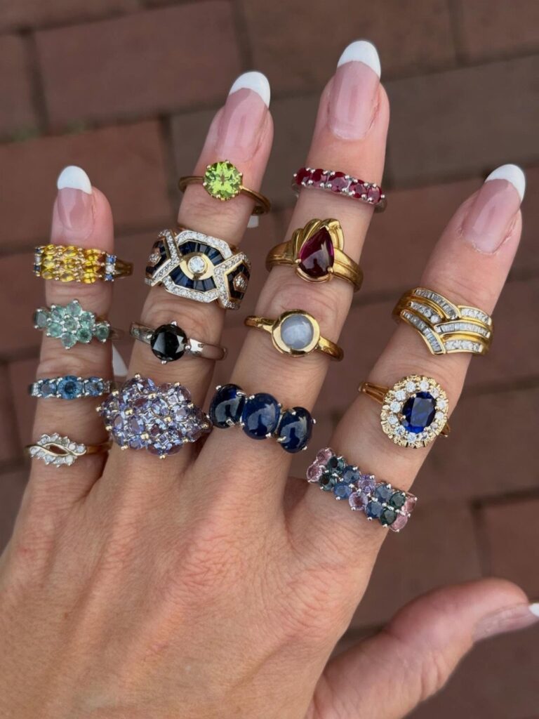 Hand wearing multiple colorful gemstone rings on every finger against a brick background