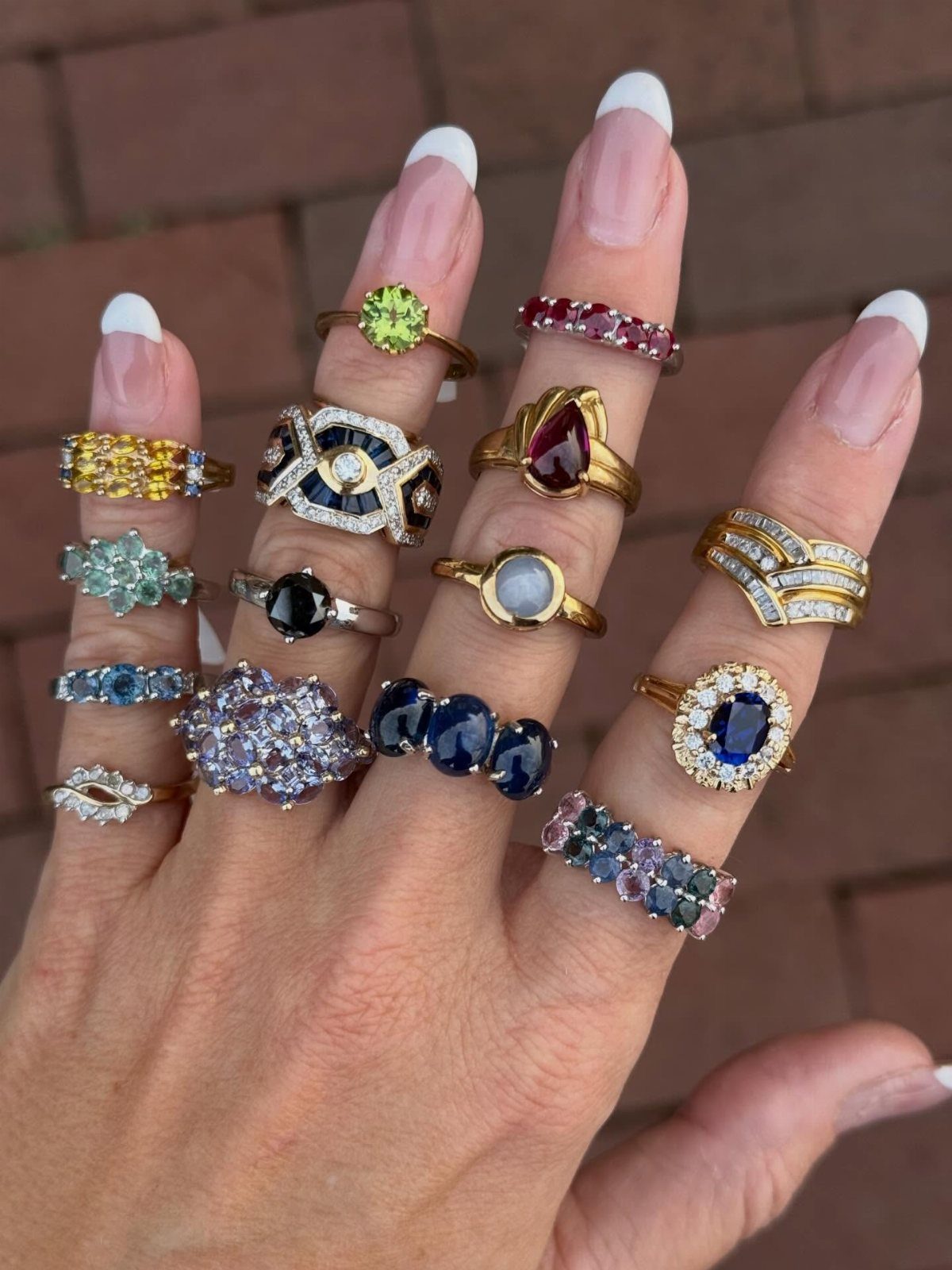 Hand wearing multiple colorful gemstone rings on every finger against a brick background
